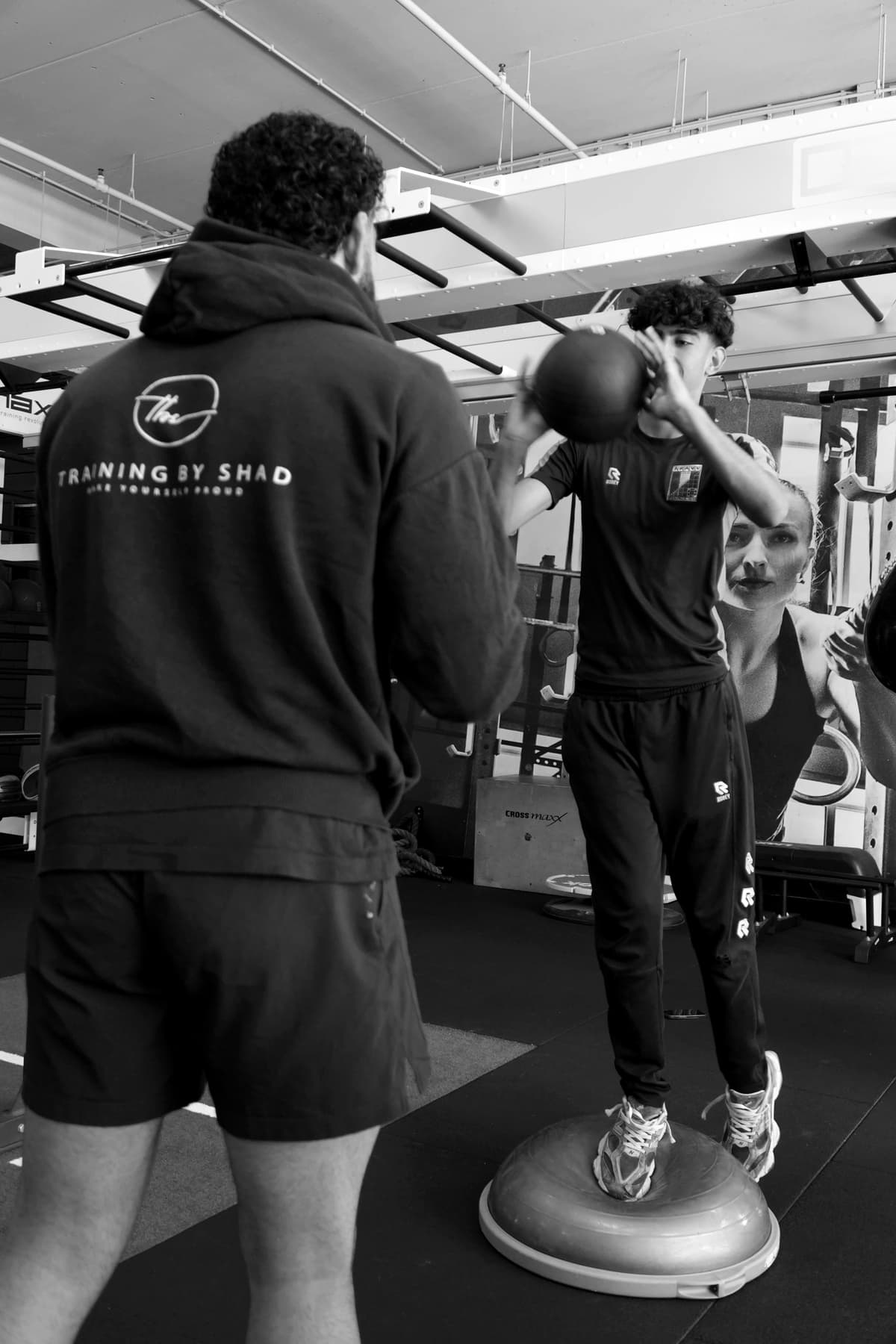 Shad, met ‘Training by Shad’ op zijn hoodie, coacht een klant die balanceert met een medicijnbal